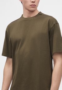 Jeune homme portant un t-shirt uni vert olive à manches courtes avec "BOSS" brodé sur l'encolure.