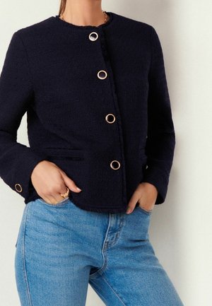 Femme portant une veste noire texturée avec des boutons en anneaux dorés et un jean bleu clair, les mains dans les poches, sur un fond clair uni.