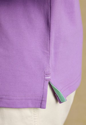 Gros plan sur l'ourlet d'un polo violet avec une bordure intérieure verte et une petite fente latérale, porté sur un pantalon beige clair.
