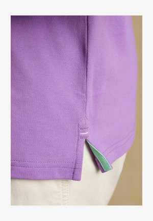 Gros plan sur l'ourlet d'un polo violet avec une bordure intérieure verte et une petite fente latérale, porté sur un pantalon beige clair.
