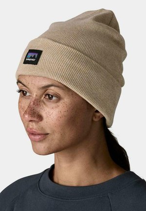 Mujer con pecas, lleva un gorro de punto beige de Patagonia y una sudadera oscura, mirando hacia la izquierda con el cabello recogido, sobre un fondo neutro.