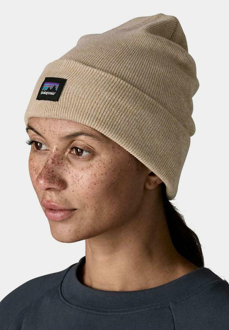 Mujer con pecas, lleva un gorro de punto beige de Patagonia y una sudadera oscura, mirando hacia la izquierda con el cabello recogido, sobre un fondo neutro.