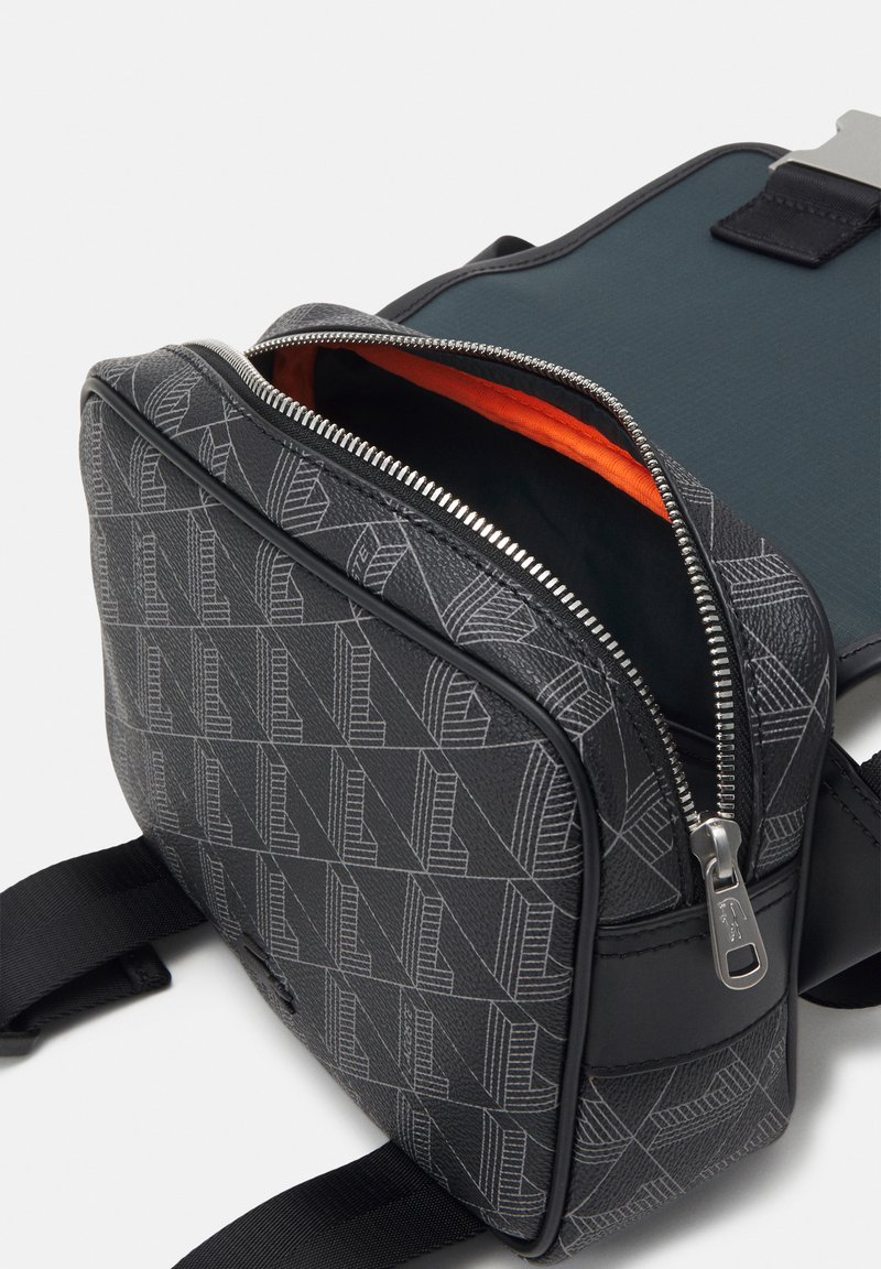 Schwarze Tasche mit geometrischem Muster, silbernem Reißverschluss, orangefarbenem Innenraum und strapazierfähigem Stoff. Verfügt über verstellbare Träger und ein elegantes Design.
