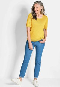 Geel kortemouwen breitop met een ronde hals, gecombineerd met blauwe tapering jeans en witte sneakers, staand tegen een lichte achtergrond.