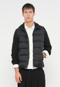 Gilet en duvet noir avec une fermeture éclair à l'avant, featuring des manches en tricot et un col haut, porté sur une chemise blanche, associé à un pantalon à carreaux.