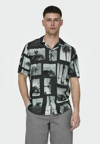Camisa de manga corta con un patrón de collage fotográfico en blanco y negro que presenta palmeras y escenas de playa. Tejido ligero con un corte relajado.