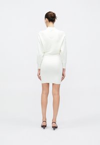 Frau mit hochgestecktem Haar, die ein weißes Langarmoberteil und einen eng anliegenden weißen Rock trägt, von hinten stehend, in schwarzen, flachen Schuhen auf weißem Hintergrund.