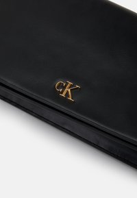 Svart lær clutch med et gull "CK"-logo foran. Glatt tekstur med en slank, rektangulær form og minimalistisk design.