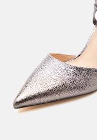 Ασημί μεταλλικό γυναικείο παπούτσι με μυτερή μύτη, ψηλό τακούνι, δίχρωμο ραβδωτό δέρμα και μπεζ εσωτερική επένδυση σε λευκό φόντο.