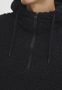Svart fleece-pullover med strukturert overflate, høy krage og halvlukking med glidelås. Har snorer i hetten og forsterkede sømmer.