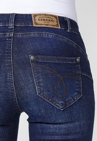 Gros plan de la poche arrière d'un jean en denim bleu foncé avec des surpiqûres décoratives et une étiquette de marque en cuir marron au-dessus de la ceinture.
