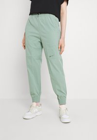 Person som bär ljusgröna Nike-joggers med elastiska manschetter och vita sneakers, stående mot en vit bakgrund.