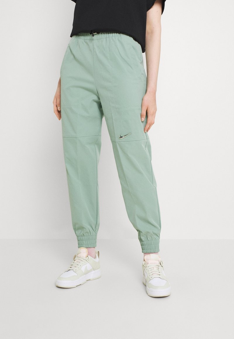 Person som bär ljusgröna Nike-joggers med elastiska manschetter och vita sneakers, stående mot en vit bakgrund.