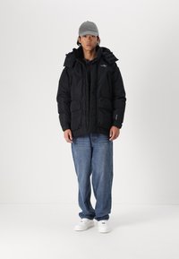 Sort pufferjakke med hætte, der har en frontlynlås og to store lommer. Bæres sammen med blå denimjeans og hvide sneakers.