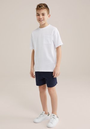 Weißes T-Shirt mit kurzen Ärmeln und einer Tasche, kombiniert mit marineblauen Shorts und weißen Sneakers, vor einem neutralen Hintergrund stehend.