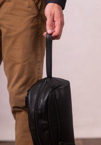 Black leather bag with a top handle, rounded shape, prominent zippers, and textured surface, held by a person wearing tan pants.
