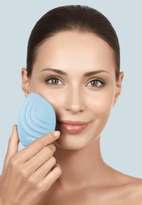 Un outil de nettoyage facial en silicone bleu avec des rainures courbes, tenu contre la joue d'une femme, présentant une texture lisse et une forme ergonomique.