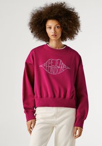 Mladá žena s bujnými kudrnatými vlasy, oblečená v magentovém svetru s logem ve tvaru rtů "Pepe Jeans London" a smetanovými kalhotami.