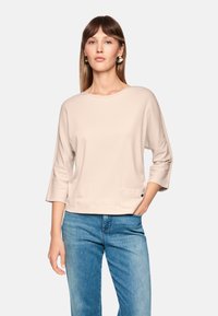 Beige 3/4 sleeve top with a round neckline and a front pocket; worn with light blue jeans, featuring a subtle texture.
