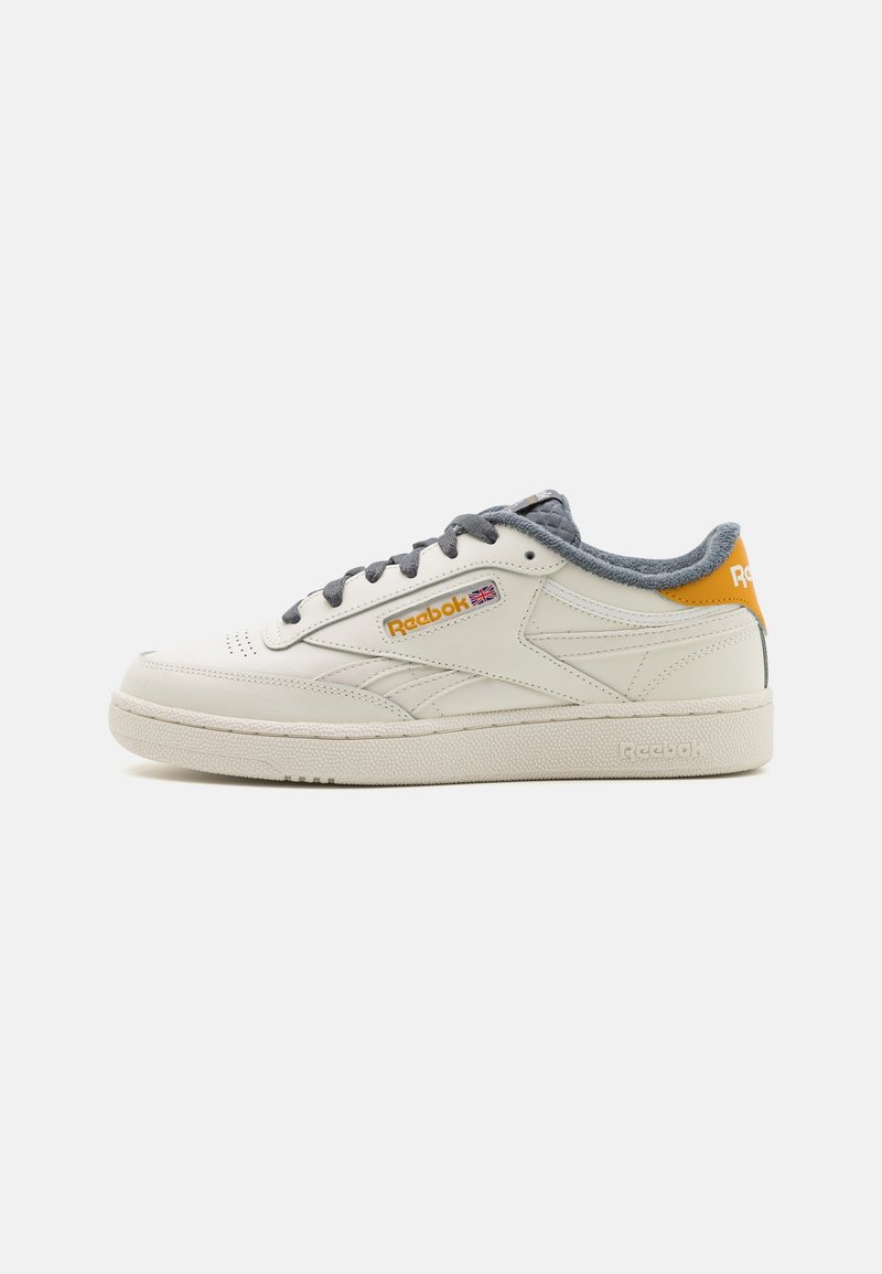 Baskets basses crème Reebok avec lacets gris, languette orange au talon et logo Union Jack sur le côté, affichées sur fond blanc.