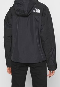 Svart jacka med huva, med en slät, syntetisk yta. Baksidan visar en vit logotyp från The North Face på övre vänstra delen.