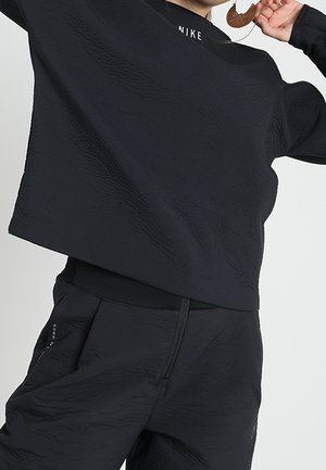 Person iført sort Nike sportstøj, inklusive en tekstureret langærmet top og matchende bukser, der holder et brunt vedhængs-halskæde.