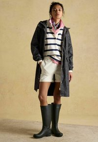 Čierny vzorovaný pršiplášť s kapucňou, biela košeľa s námorníckymi pruhmi, smotanové šortky a zelené gumové topánky po kolená. Ružová šatka ako akcent.