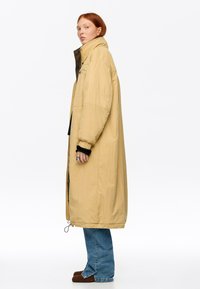 Mujer pelirroja con un abrigo largo acolchado beige, jeans azules y zapatos marrones, de pie de perfil contra un fondo blanco.