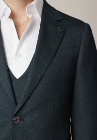 Mężczyzna w ciemnogranatowej marynarce o fakturowanej powierzchni z przypinką na klapie i białej koszuli, rozpiętej pod szyją, z widocznymi owłosieniami na klatce piersiowej.