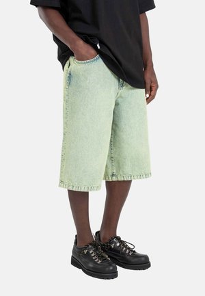 Person, die hellgrüne lange Jeansshorts, ein schwarzes oversized T-Shirt und schwarze Schnürschuhe vor einfachem Hintergrund trägt.