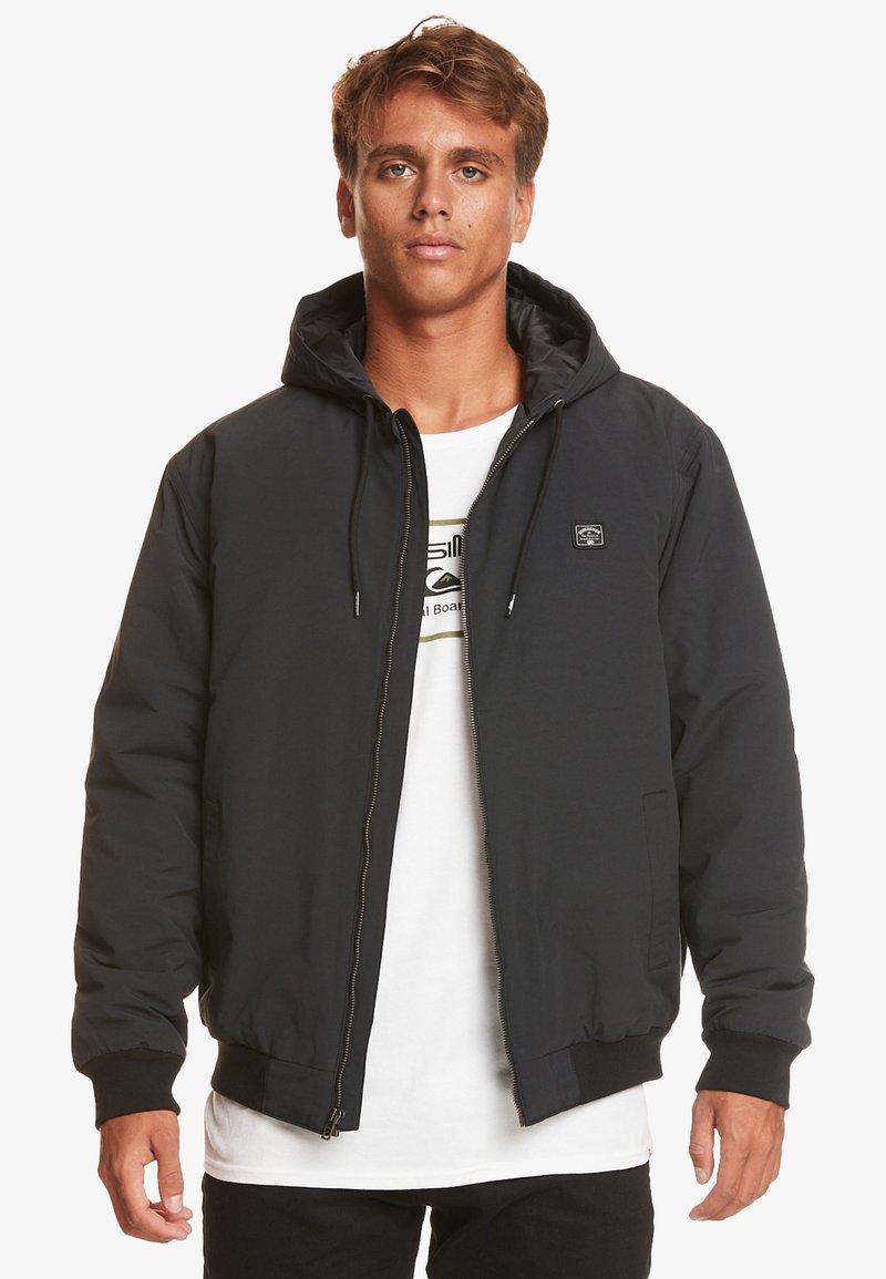 Black padded jacket with a hood, front zipper, and ribbed cuffs. Features a small logo patch on the chest. Worn over a white shirt.