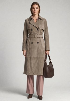 Kvinde iført taupe trenchcoat med bælte og lyserøde bukser, der holder en mørkebrun lædertaske, står foran en neutral baggrund.