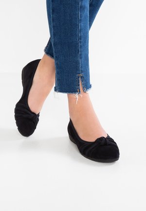 Kvinde iført ankellange blå jeans med rå kanter og sorte ruskindssko med knudedetalje på glat hvidt gulv.