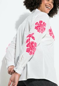 Weißes Hemd mit Knopfverschluss und großem, plüschigem, pinkfarbigen floralem Stickdesign an beiden Ärmeln. Mit Knopfmanschetten und lockerer Passform.