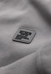 Šedá látka se hladkým povrchem obsahuje černou čtvercovou etiketu s embosovaným textem včetně "SINCE 1983" a zakřiveným okrajem.