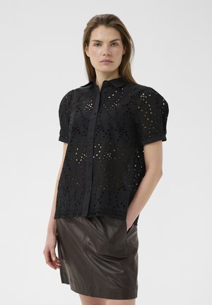 Frau trägt eine schwarze Bluse mit kurzen Ärmeln aus Lochspitze und gepufften Schultern sowie einen dunkelbraunen Lederrock, steht mit einer Hand in der Tasche.