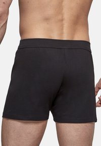 Miehen torso ja yläjalat, jotka käyttävät yksinkertaisia mustia bokserihousuja, näkyvät takaa valkoista taustaa vasten.