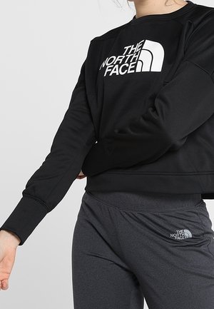 Personne portant un sweat-shirt noir et un pantalon gris foncé, tous deux avec des logos blancs The North Face, montrée du cou aux hanches.