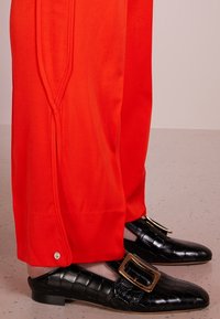 Jasnoczerwone spodnie z bocznymi szwami i napem u dołu, zestawione z czarnymi loafersami o fakturze krokodylej, które mają dużą z�łotą klamrę.