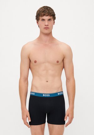 Jovem com cabelo curto e encaracolado, vestindo boxers pretos com uma faixa azul rotulada "BOSS", de pé contra um fundo branco simples.