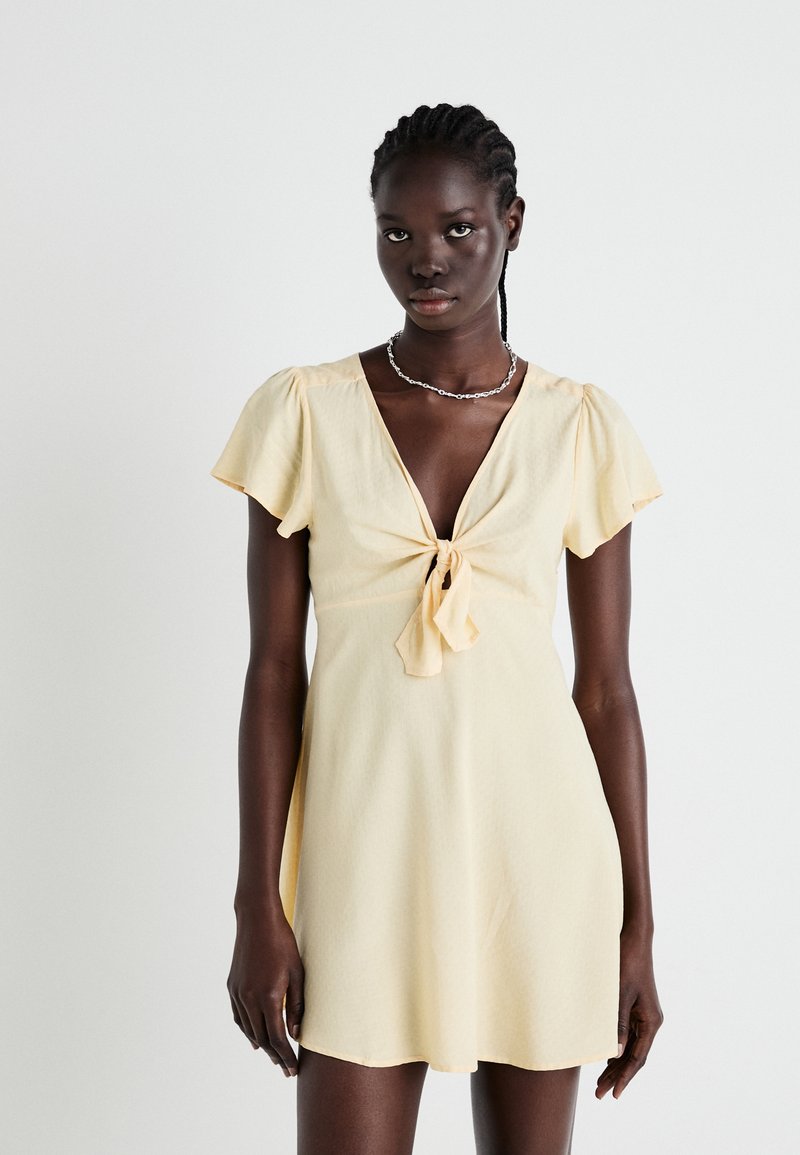 Short yellow dress with a deep V-neck, tie detail at the center, and short flutter sleeves. The fabric has a subtle texture.
