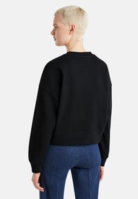 Černá zkrácená mikina s kulatým výstřihem, spadlými rameny a žebrovaným lemem. Modelka má na sobě modré kalhoty, pohled zezadu.