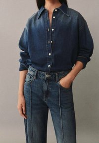 Camisa de mezclilla en azul oscuro con cuello, botones en la parte delantera y mangas largas. Combinada con jeans de tiro alto en azul oscuro con piernas rectas y costuras visibles.