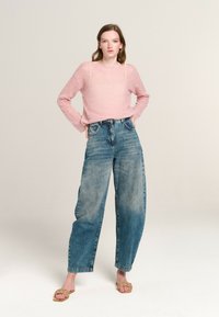 Helles, rosa, strukturiertes Strickoberteil kombiniert mit hoch taillierten, ausgebleichten, weiten Jeans und beigefarbenen, geflochtenen Sandalen. Das Model posiert vor einem neutralen Hintergrund.