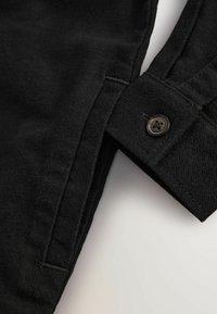 Schwarze knöpfbare Bluse mit gewebter Textur, ausgestattet mit einem dunklen Knopf und sauberem Stich entlang des Randes und der Manschette.