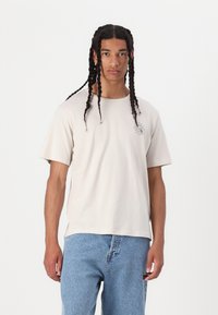 Tricou bej cu mânecă scurtă, fabricat dintr-un material moale, având un grafic cu soare pe partea stângă a pieptului, asortat cu blugi din denim de culoare albastru deschis.