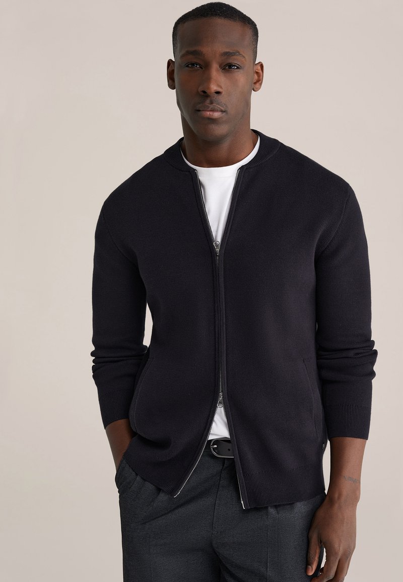 Tamsiai mėlynas užsegamas kardiganas su apvalia apykakle, apsiūtais rankogaliais ir apačia, vilkimas virš baltos marškinėlių, derinamas su pilkomis kelnėmis. Lygios tekstūros.