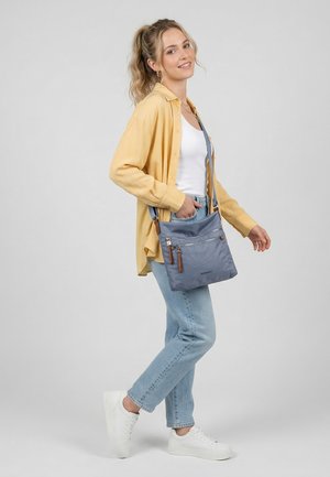 Junge Frau lächelnd, trägt gelbes Hemd, weißes Oberteil, hellblaue Jeans, weiße Turnschuhe, hält eine graue Umhängetasche vor einfarbigem Hintergrund.