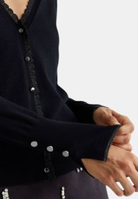 Cardigan nero con rifiniture in pizzo scallop lungo il colletto, bottoni argentati sui polsini e una texture a coste.