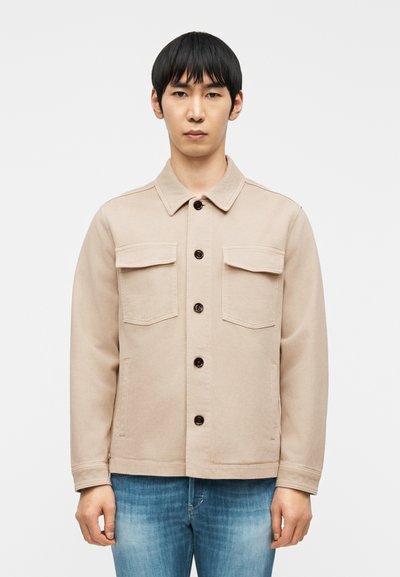 Jeune homme aux cheveux noirs lisses portant une veste beige boutonnée avec des poches poitrine et un jean bleu, debout devant un fond uni.